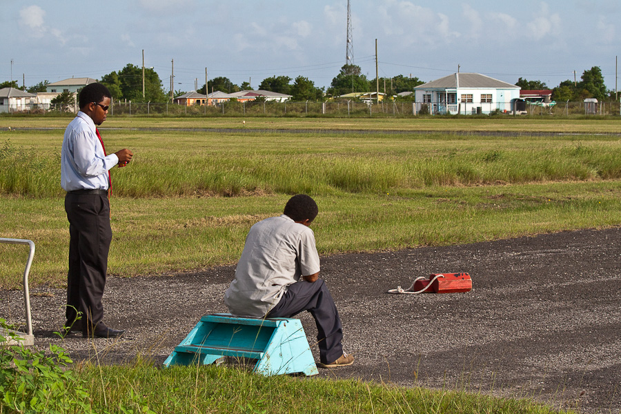 Два чёрных мужчины-работника аэропорта в тёмных очках ожидают рейса на лётном поле. Аэропорт Барбуда Кодрингтон, остров Барбуда, Антигуа и Барбуда. Two black male airport workers in dark glasses are waiting for a flight on an airfield. Barbuda Codrington Airport (IATA: BBQ), Barbuda island, Antigua and Barbuda, Caribbean.