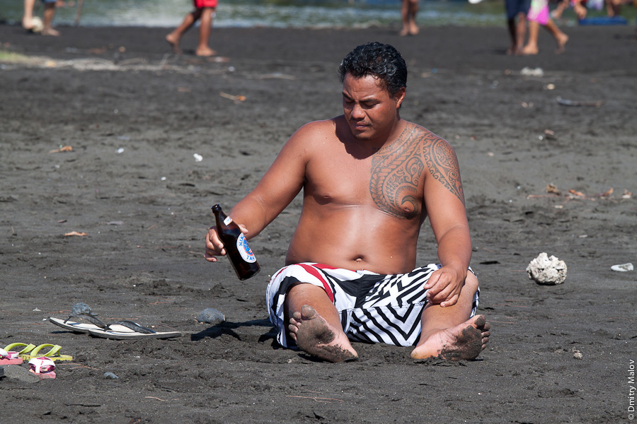 Venus beach, Pointe Venus, Tahiti, French Polynesia. Пляж Пойнте-Венус, Таити, Французская полинезия, пляж с чёрным песком, полинезиец-мужчина с полинезийской таитянской татуировкой сидит с пивом Хинано. A tattooed polynesian guy sits on a black sandy beach with tahitian Beer Hinano