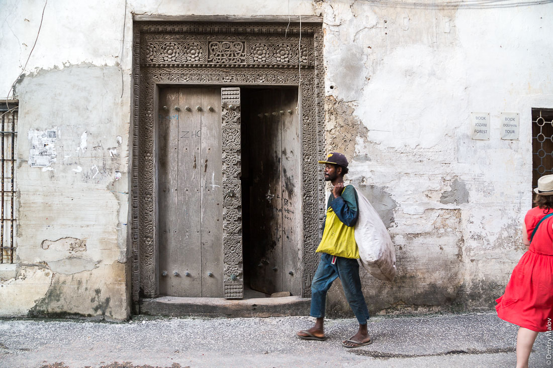 Чёрный мужчина с мешком и женщина в красном платье. Резные занзибарские двери. Каменный город, Занзибар-сити, остров Унгуджа, Танзания. A black man with a bag and a woman in a red dress. Carved Zanzibar doors. Stone Town, old town of Zanzibar City, Unguja island, Tanzania.  