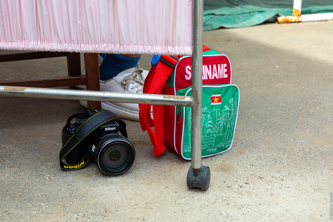 Фотооаппарат Nikon и рюкзак Suriname лежат на земле пока туристу за ширмой берут тест на COVID-19, Сан-Томе и Принсипи